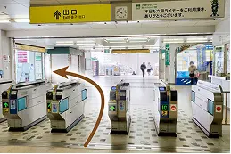自動改札を出た先の通路。頭上に黄色い「出口」の案内板があり、左方向へオレンジ色の矢印が示されている。
