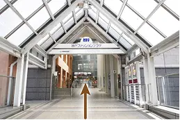 ガラス屋根の連絡通路。奥に「神戸ファッションプラザ」の案内板が見え、通路の先へ進む方向にオレンジ色の矢印が示されている。