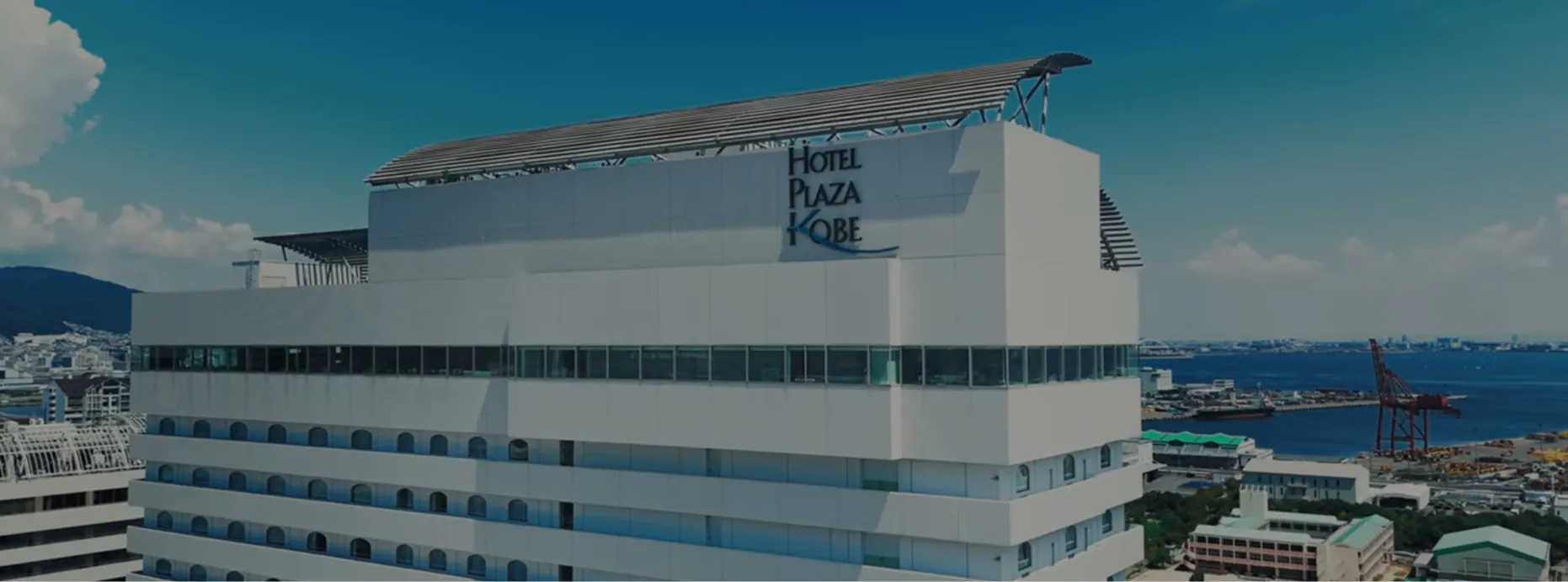 青空を背景に立つホテルプラザ神戸の外観。屋上に「HOTEL PLAZA KOBE」のロゴがあり、奥には神戸港と海が広がっている。
