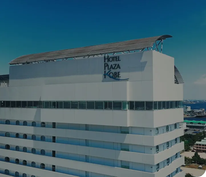 青空を背景に立つホテルプラザ神戸の外観。屋上に「HOTEL PLAZA KOBE」のロゴがあり、奥には神戸港と海が広がっている。