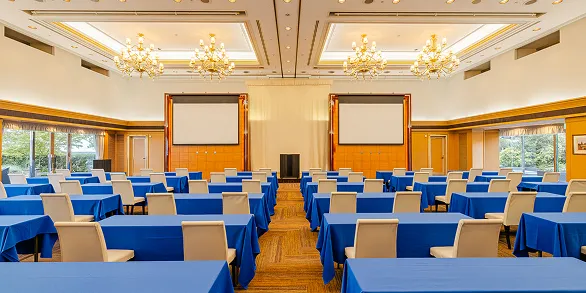 スクール形式に設営した宴会場。青いクロスの長机が中央の通路を挟んで左右のブロックに分かれて並び、正面にはスクリーンが2面、天井にはシャンデリアが並んでいる。
