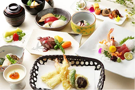 黒い竹かごに盛られた海老や野菜、椎茸の天婦羅を中心に、海老や鮪のお造り、牛肉料理、焼き物、茶碗蒸し、蓋付きの椀などが、和の器に彩りよく盛り付けられて並ぶ写真。