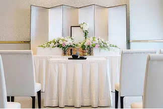 白銀の屏風を背にした祭壇に、写真立てと白やピンクの花が横長に飾られた様子。祭壇は白いひだ飾りのクロスで覆われ、中央に香炉が置かれている。両脇には白い椅子が並ぶ。