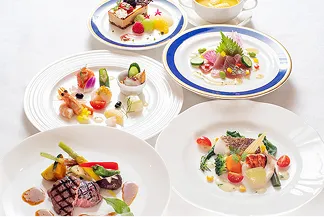 青い縁取りの皿と白い大皿に盛り付けられたコース料理が、白いテーブルの上に並ぶ写真。牛フィレ肉のグリルと彩り野菜、魚のポワレ、前菜の盛り合わせ、カルパッチョ、スープ、デザートが見える。