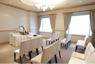 大きな窓から明るい光が差し込む会場。左奥に花で飾られた祭壇と焼香台が設けられ、その前に白い椅子が中央の通路を挟んで何列も並ぶ。天井にはシャンデリアが下がっている。