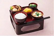 黒塗りの脚付きのお膳に、朱色の器に盛った白いご飯と汁物、煮物、野菜の和え物、香の物などの小鉢が並び、手前に箸が添えられた写真。仏前にお供えするための一膳が整えられている。