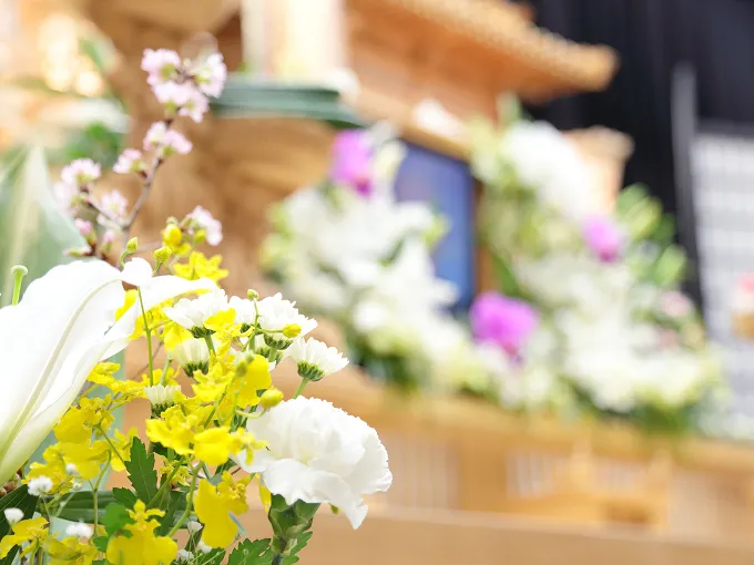 白や紫の花で飾られた木目調の祭壇を背景に、手前に白いユリやカーネーション、黄色い小花、ピンクの花を取り合わせた供花が写る写真。奥の祭壇はやわらかくぼかされ、花の彩りが際立っている。