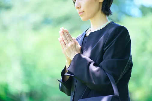 明るい緑の木々を背景に、黒い喪服に真珠のネックレスを身につけた女性が、目を伏せて胸の前で静かに手を合わせている様子。やわらかな自然光の中、穏やかに祈りを捧げる姿を写した写真。