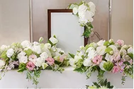 白い写真立てを中心に、その両脇と上部を白やピンクの花と緑の葉で横長に華やかに飾った祭壇装花の写真。やわらかな色合いの花が写真立てを包み込むように生けられている。