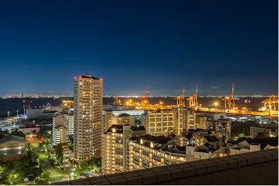高層階から見下ろした夜景の写真。明かりの灯る高層マンションや建物の向こうに、オレンジ色の光に照らされた港のクレーン群と、海の対岸に連なる街の灯りが広がっている。