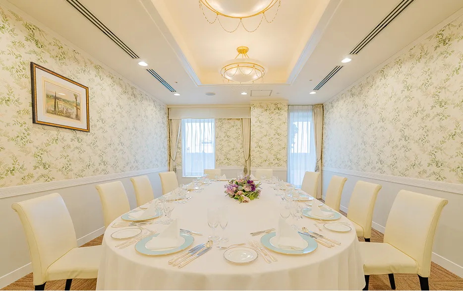 小宴会場「沖・港」の会食スタイル。白いクロスの楕円形テーブルの中央に紫とピンクの花が飾られ、アイボリーの椅子が囲んでいる。壁は緑の草花柄の壁紙で、天井にはシャンデリア、奥にはレースカーテンの窓がある。