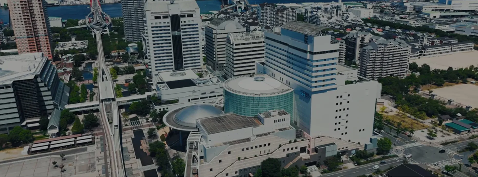 神戸の街並みを上空から見下ろした風景。高層ビルが立ち並ぶ中、モノレールの高架線とドーム状の建物が並ぶ一角にホテルプラザ神戸が建っている。