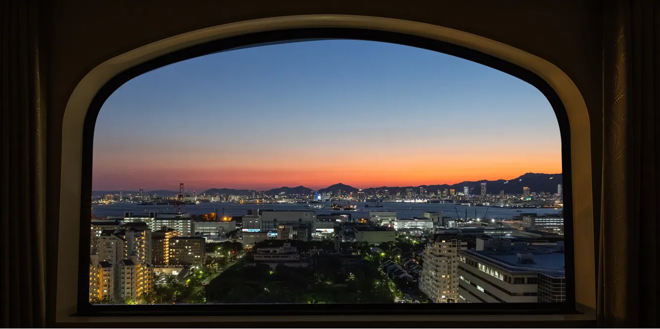 アーチ型の窓から見える夕暮れの神戸港。オレンジと青のグラデーションに染まる空の下、明かりがともり始めた港の建物群と山並みのシルエットが広がる。