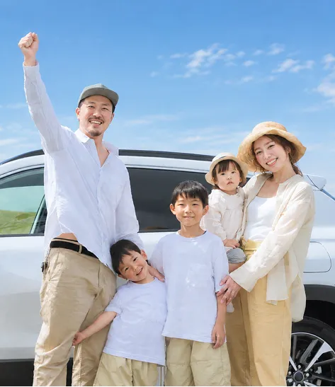 白い車の前で笑顔を見せる家族。ガッツポーズをする父親と、子どもたちを抱っこする母親が並んで立っている。
