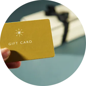 金色の「GIFT CARD」と書かれたカードを持つ手。背景には黒いリボンがかかったギフトボックスが置かれている。