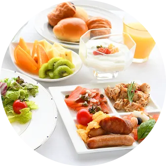 ホテルの朝食ブッフェ料理。ソーセージやスクランブルエッグ、サーモンを盛ったプレート、サラダ、フレッシュフルーツ、パン、ヨーグルトがテーブルに並んでいる。