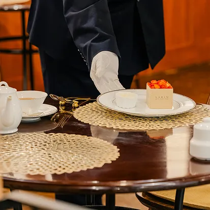 黒い制服に白い手袋のスタッフが、丸テーブルの上にいちごをのせたケーキの箱とデザートの皿を並べている。紅茶ポットも置かれたカフェの一場面。