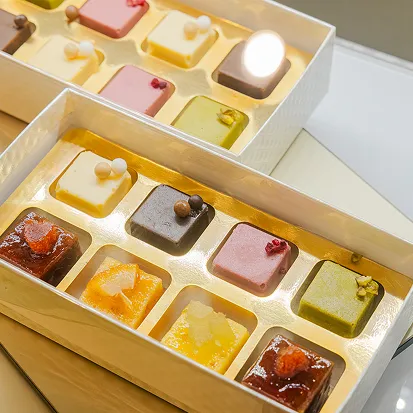 開いた白い箱の中に、いちご・抹茶・チョコレートなど彩り豊かな一口サイズのケーキが等間隔に並び、やわらかな照明に照らされている。