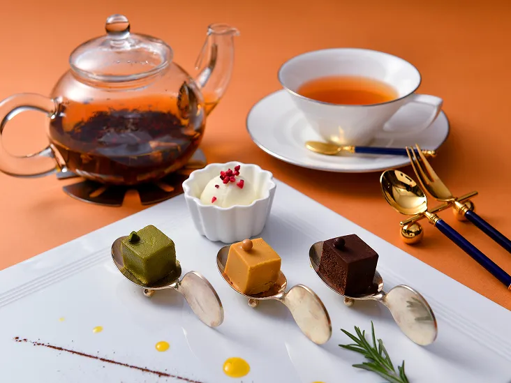 3段のケーキスタンドに、オレンジ色や抹茶、いちごをあしらった一口サイズのケーキが並び、そばにガラス製の紅茶ポットが添えられている。
