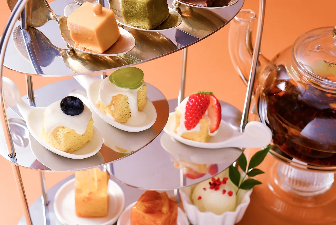 開いた白い化粧箱の中に、いちご・抹茶・チョコレートなど彩り豊かな正方形のケーキが等間隔に並び、箱の内側が金色の光でやわらかく照らされている。