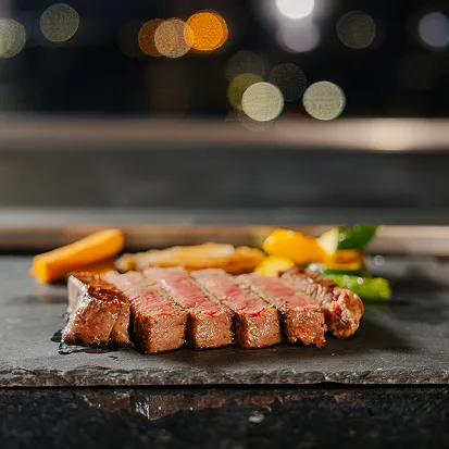 黒い石板の上に、断面がきれいな霜降り牛肉の切り身が並べられ、奥には彩り野菜が添えられている。背景には店内の明かりがぼかされて見える。