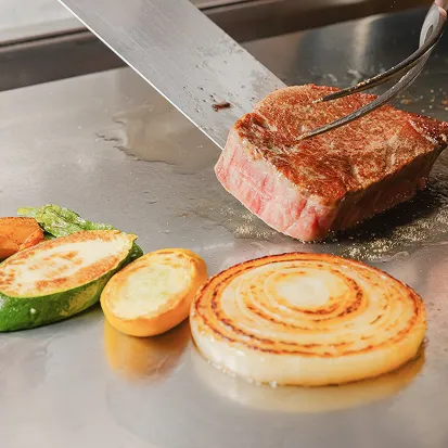 鉄板の上で、シェフがトングで牛肉を持ち上げながら、隣でズッキーニや玉ねぎの輪切りを焼いている。