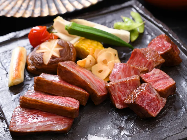 黒い石板に盛られた霜降り牛肉の切り身と彩り野菜のグリル、背景には扇子や伊勢海老の皿がぼかされて写っている。