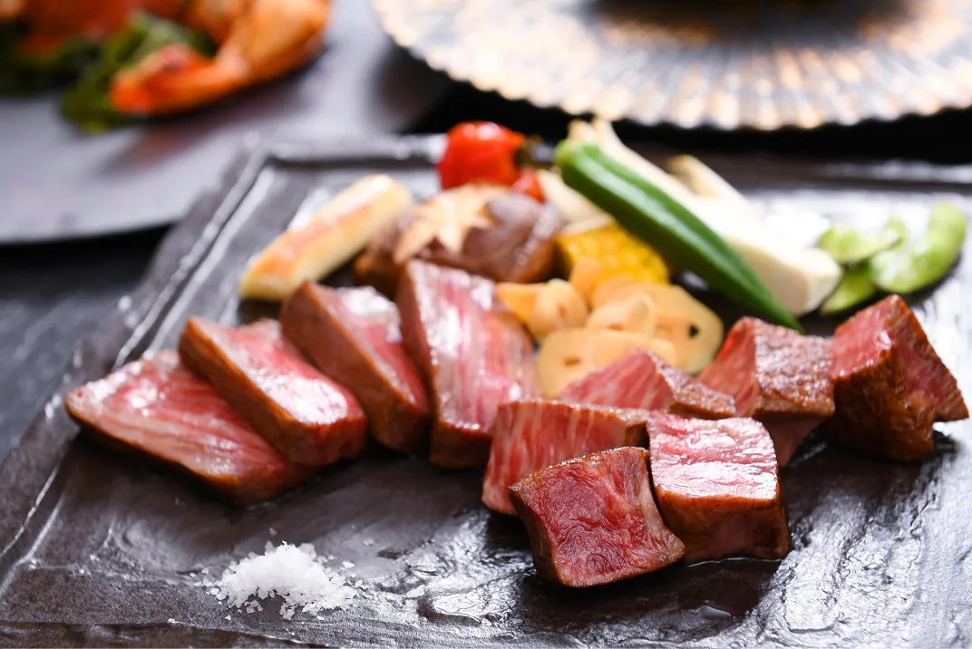 黒い石板に、断面が鮮やかな霜降り牛肉の切り身と、ミニトマトやオクラ、とうもろこしなど彩り野菜のグリルが盛りつけられ、塩が添えられている。