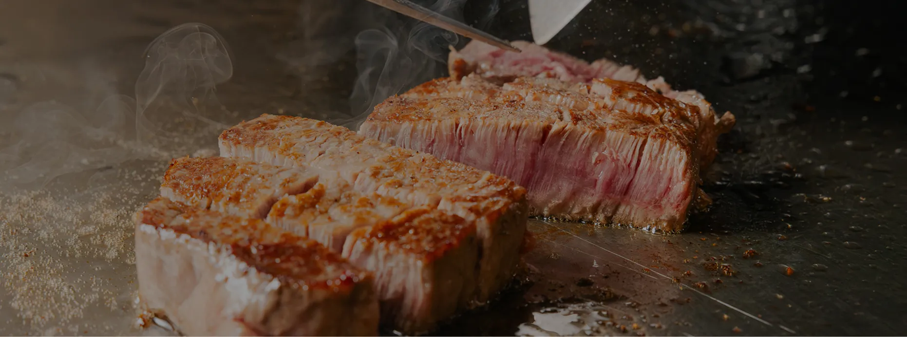鉄板の上で牛肉のステーキが焼かれ、包丁とヘラを持つシェフの手元から湯気が立ちのぼっている。