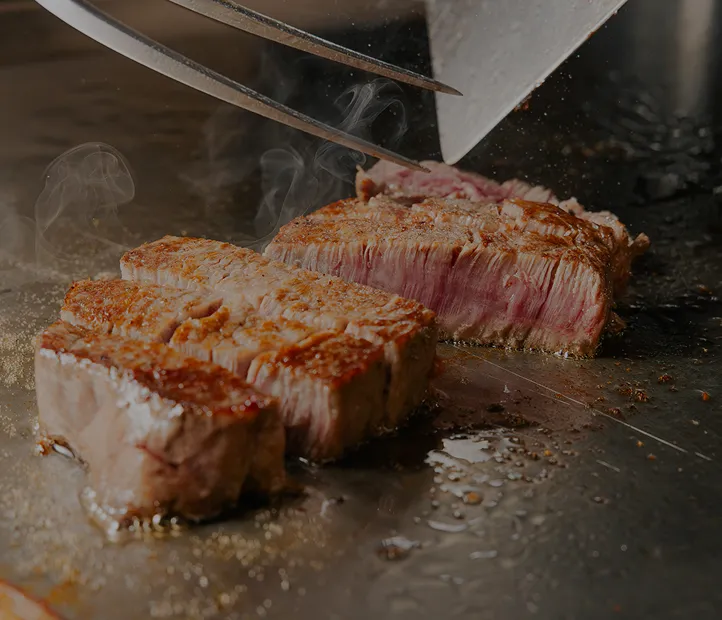 鉄板の上で牛肉のステーキが焼かれ、包丁とヘラを持つシェフの手元から湯気が立ちのぼっている。