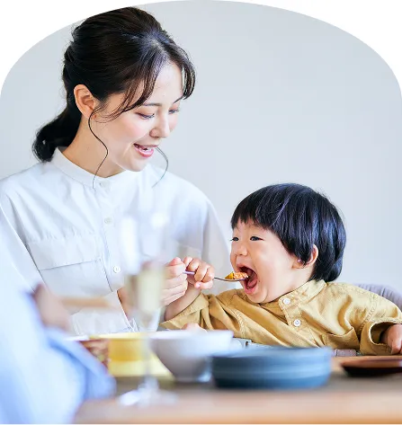 母親が笑顔でスプーンを差し出し、子どもが大きく口を開けて食事を楽しんでいる様子。