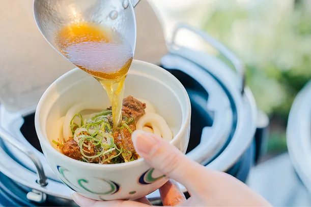 うどんと牛すじが入った丼に青ねぎと生姜をのせ、湯気の立つだし汁を注いでいる様子。