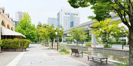 緑の街路樹が並ぶ石畳の遊歩道。水路沿いにベンチが置かれ、奥には高層ビルやモノレールの高架が見える六甲アイランドの街並み
