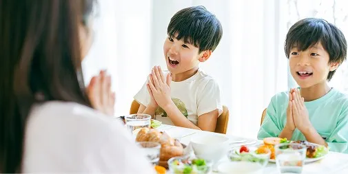 パンやサラダが並ぶ食卓を前に、笑顔で手を合わせる2人の男の子と、向かいに座る女性の後ろ姿