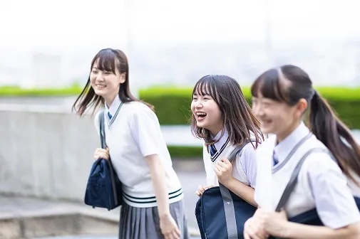 笑い合いながら歩く、制服姿の女子高校生3人。鞄を肩にかけ、街並みを見下ろす屋上のような開けた場所を歩いている。