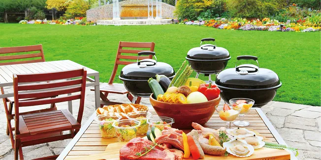 赤いガーデンチェアが並ぶテラスに、黒いBBQコンロと肉・野菜、前菜を並べたテーブル。奥には芝生と花壇が広がる。