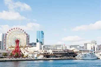青空の下、海沿いに赤い観覧車や商業施設が並び、岸壁に白い客船が停泊するハーバーランドの風景