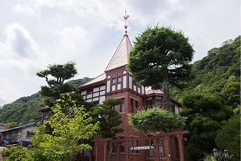 緑の木々と山を背に、尖塔と風見鶏を載せた赤レンガ造りの洋館が建つ北野異人館街の風景