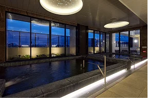 夜景が見える大浴場。湯船の奥の大きな窓ガラス越しに街の夜景が広がっている。