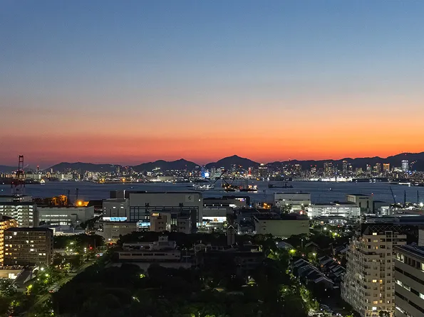 夕暮れ時の神戸の街並みと海。山のシルエットとともに、オレンジ色に染まる空と灯り始めた街の明かりが広がる。