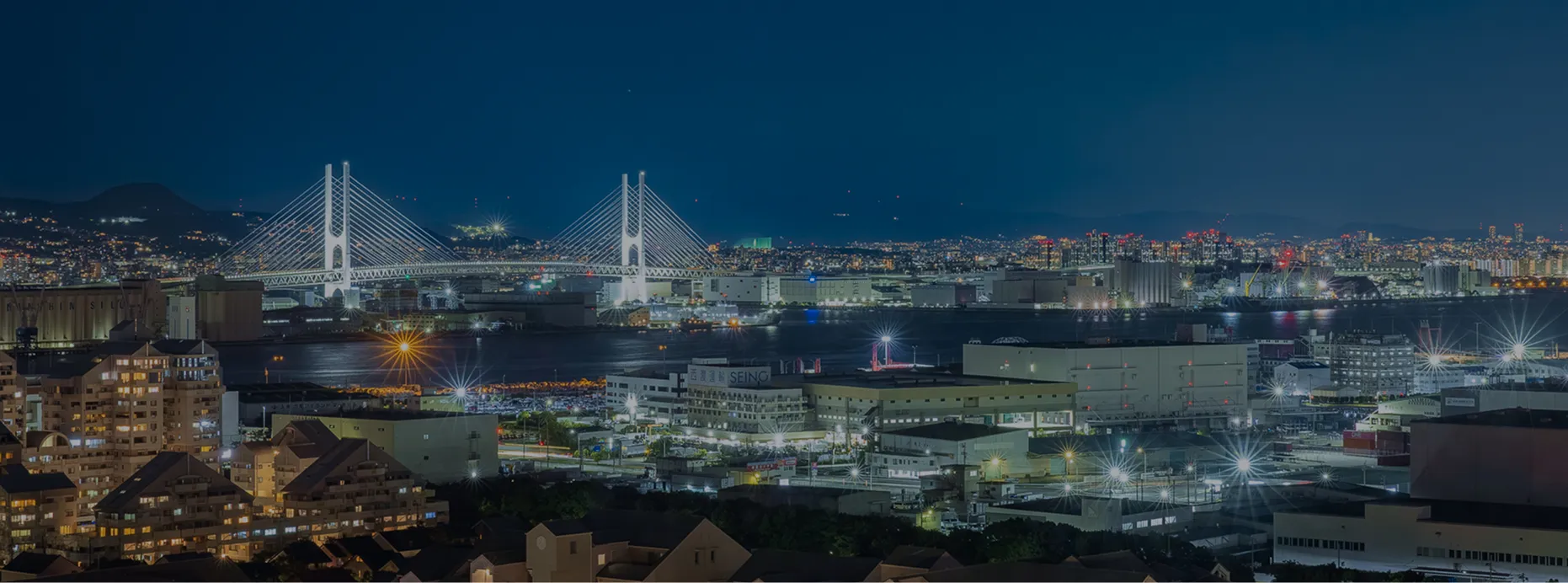 夜の神戸の街並みと、ライトアップされた斜張橋。港に明かりが映り込み、高層マンション群と山並みのシルエットが広がる。
