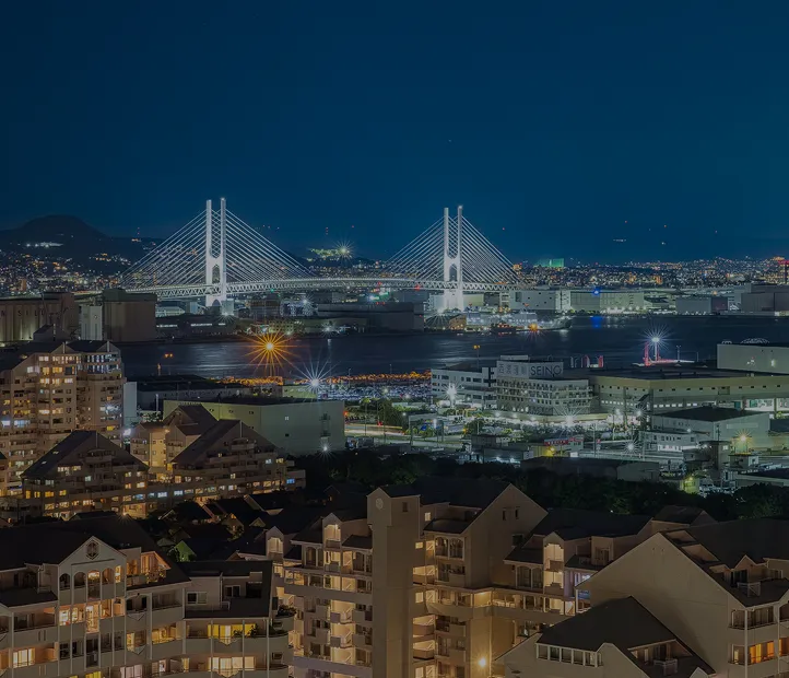 夜の神戸の街並みと、ライトアップされた斜張橋。港に明かりが映り込み、高層マンション群と山並みのシルエットが広がる。