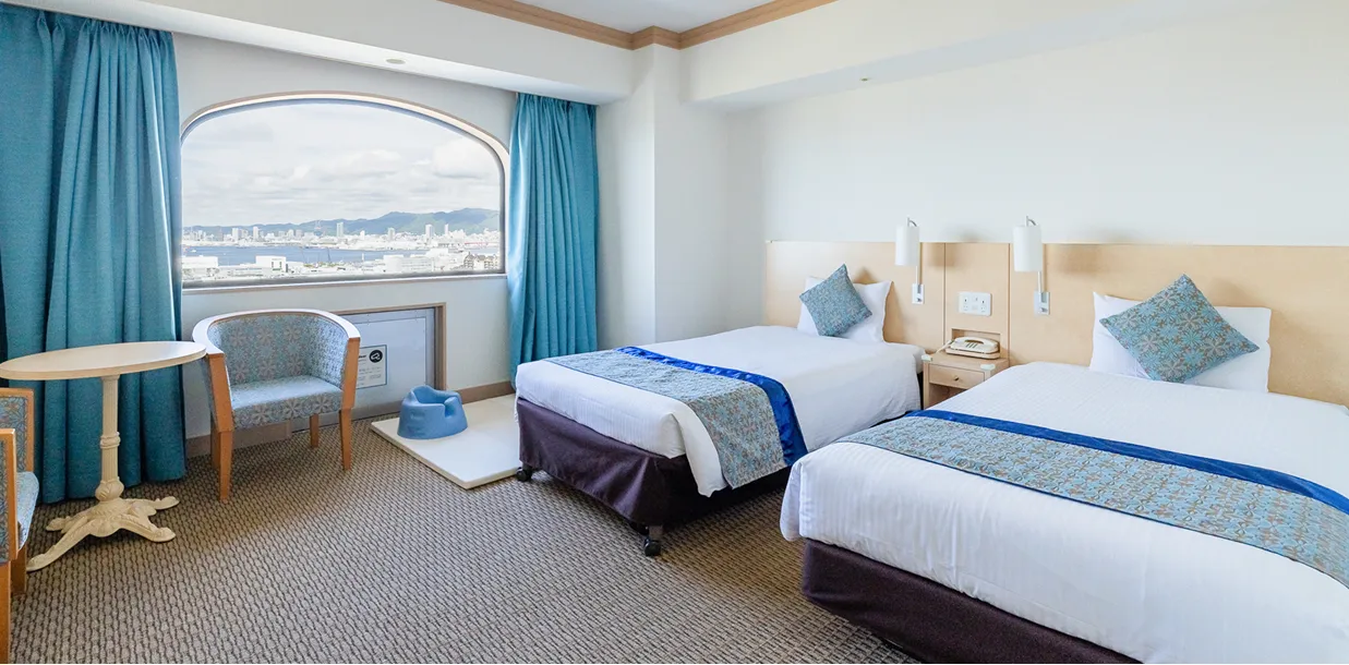 ターコイズブルーのカーテンと海の景色が印象的なツインルーム。窓際にはベビー用のおまるが置かれ、家族での利用に配慮されている。